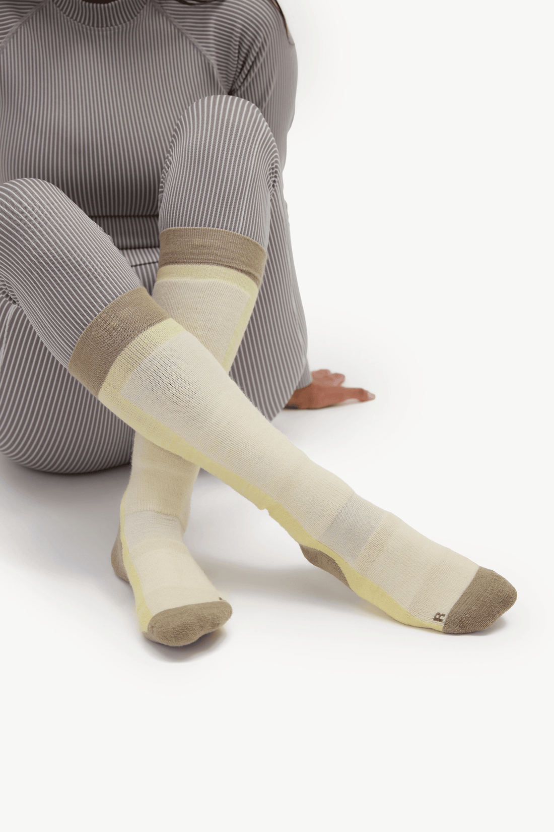 A person seated against a white studio background wearing cream and beige knee-high socks with a ribbed gray top. The socks have a pale yellow inner section and tan toe and cuff areas.