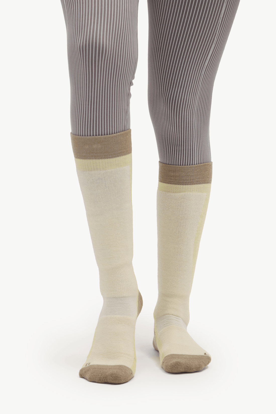 A pair of knee-high compression socks in beige and pale yellow, worn with gray ribbed tights against a white studio background.
