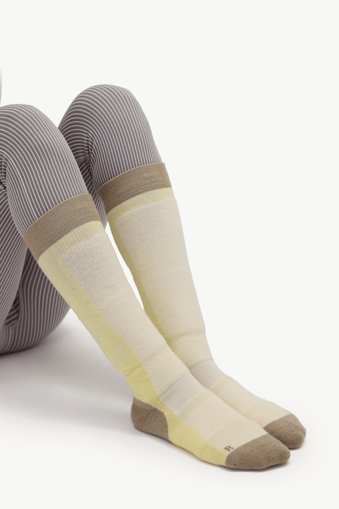 A pair of knee-high socks worn on legs, featuring a color-blocked design with gray and white vertical stripes on the upper leg, a light beige band, a pale yellow section, and taupe toes and heels, photographed against a clean white background.