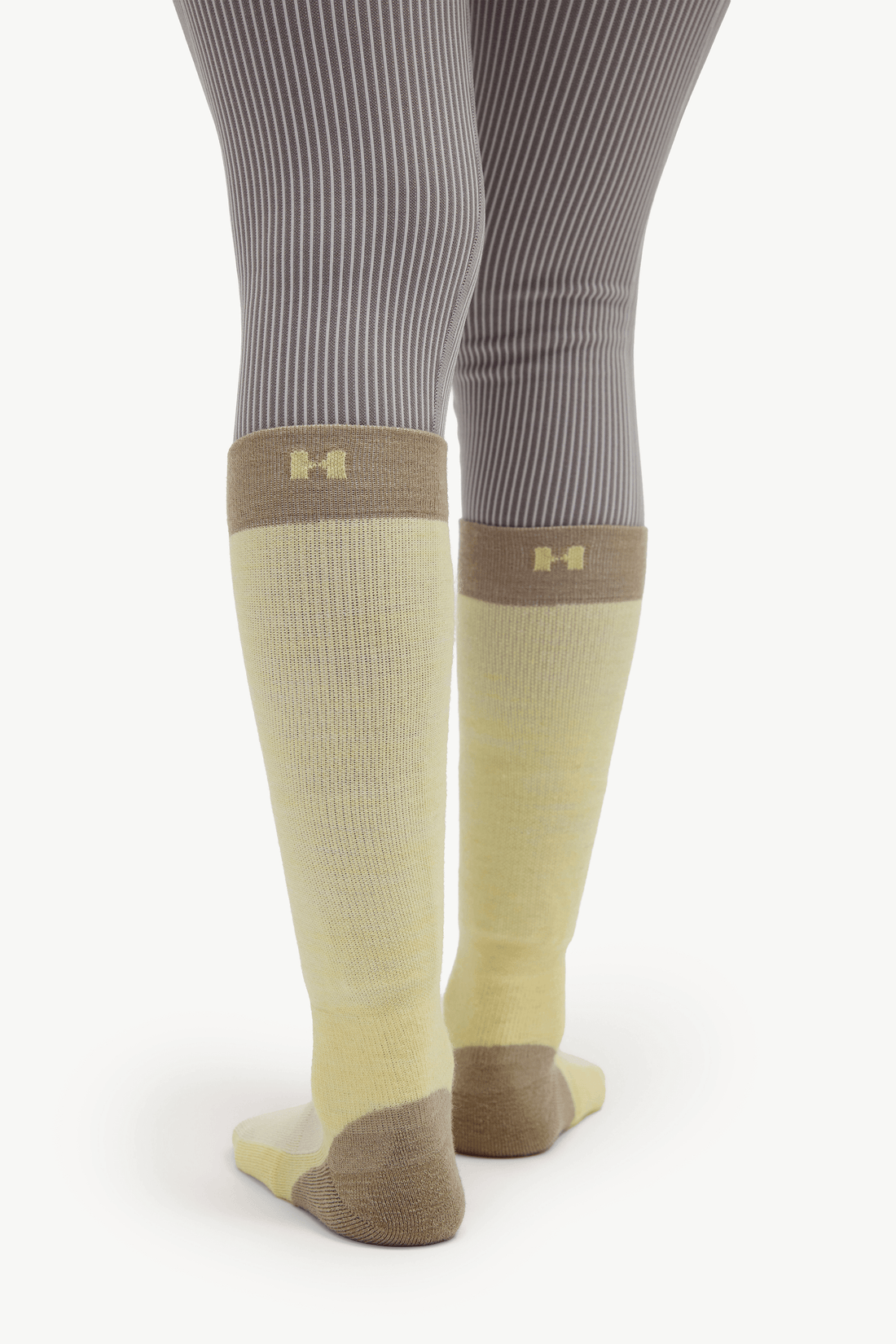 A pair of knee-high athletic socks in pale yellow and taupe, worn with gray ribbed tights. The socks feature a small gold Under Armour logo near the top cuff.