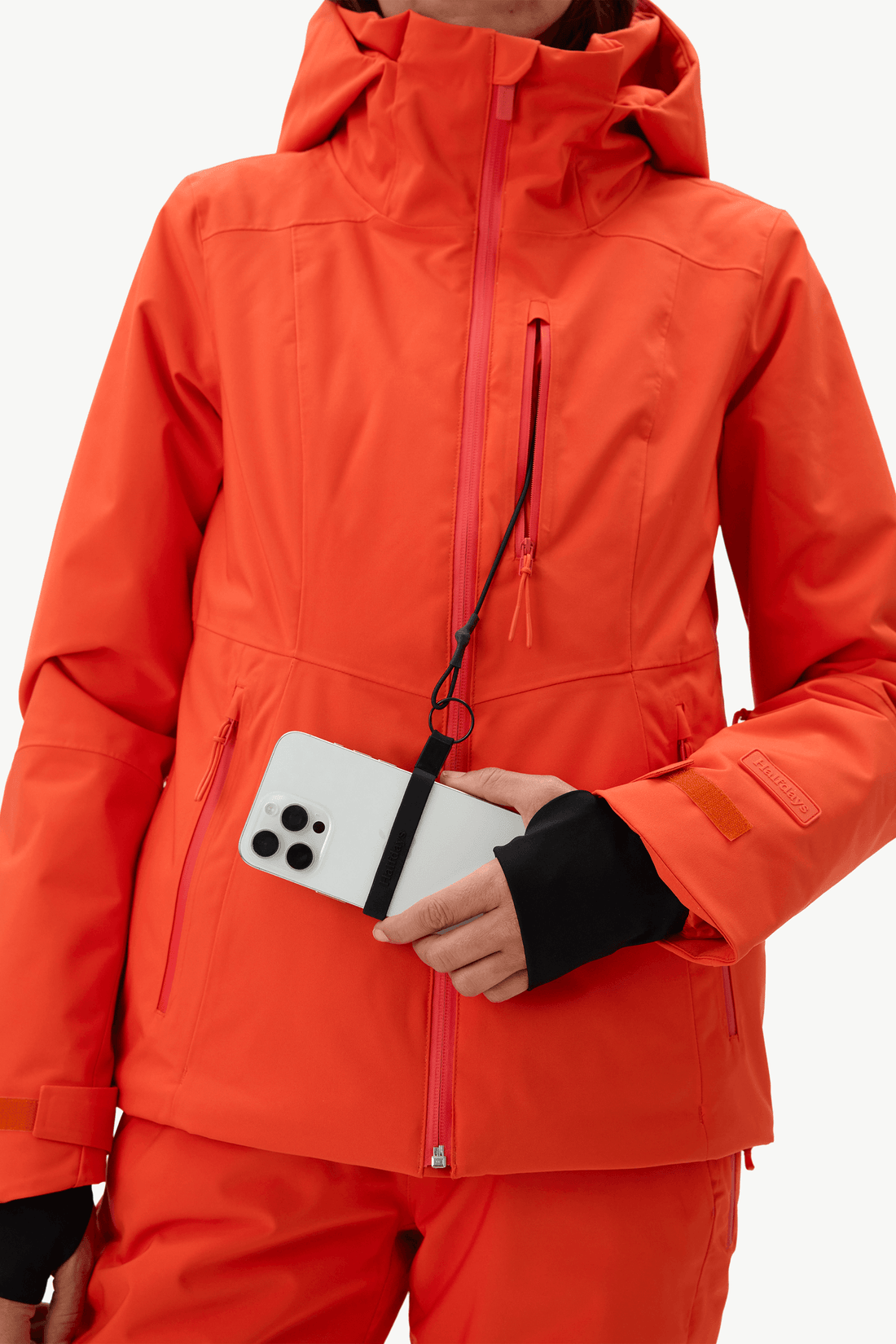 A close-up of a person wearing a bright orange ski jacket and matching pants, holding a white smartphone in a front pocket. The jacket has a high collar, a front zipper, and zippered pockets, with black wrist gaiters visible.