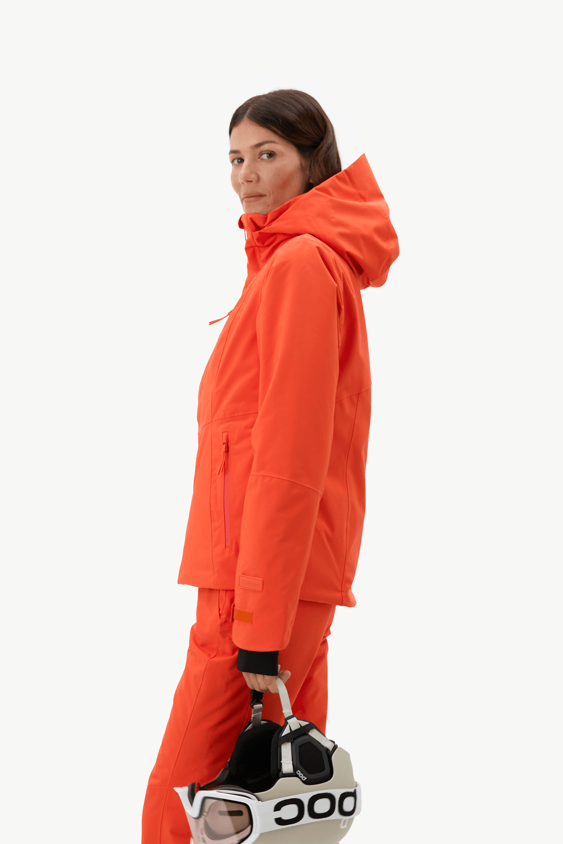 A woman in a bright orange ski jacket and matching pants stands in profile against a light gray studio background, holding a white ski helmet with black branding.