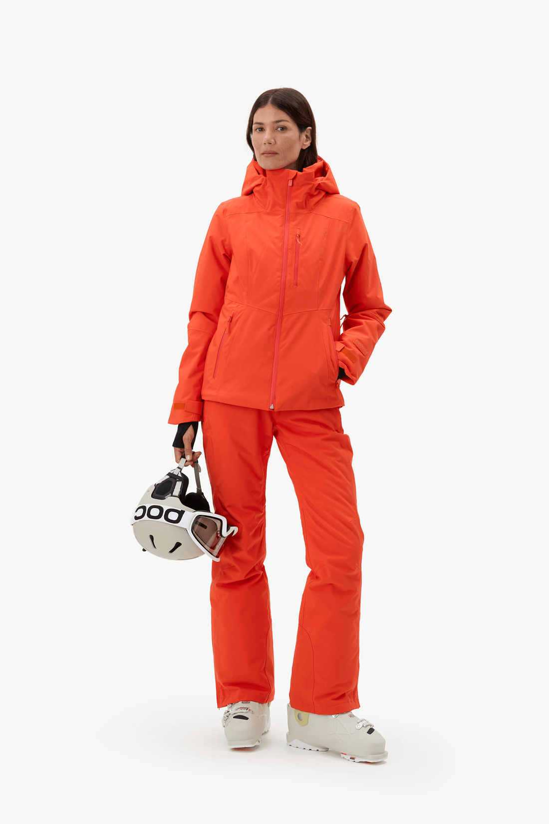 A woman modeling a bright orange ski jacket and matching ski pants, holding a white and black ski helmet against a clean white studio background.