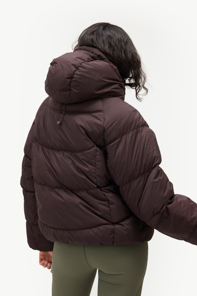 A person shown from the back wearing a dark brown puffer jacket with horizontal quilting and a hood, paired with olive green leggings, against a plain white studio background.