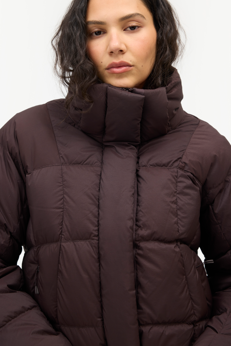 A close-up of a model wearing a dark brown puffer jacket with a high padded collar, shown against a plain light gray background.
