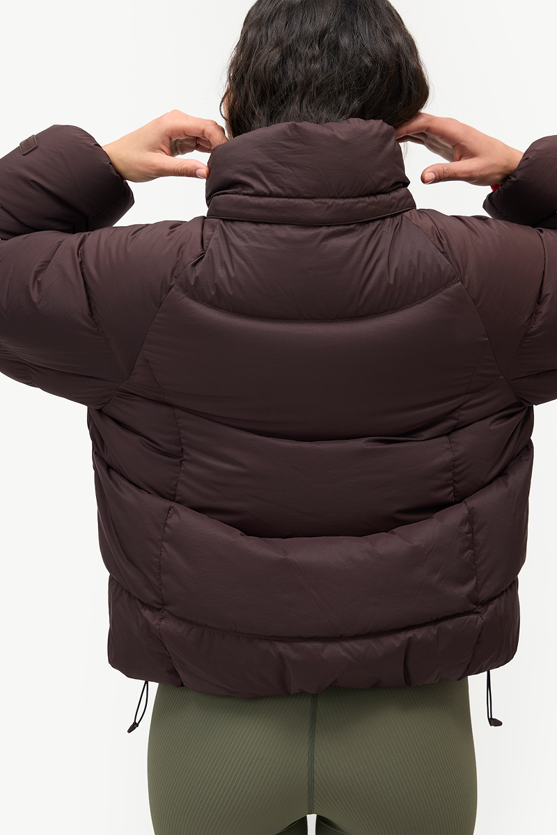 A person viewed from behind wearing a dark brown puffer jacket with a high padded collar and olive green leggings, against a plain light background.