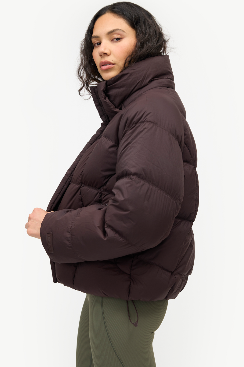 A woman wearing a dark brown puffer jacket with a high collar, shown in a side profile against a plain white studio background.