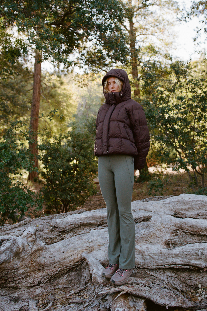 A woman stands outdoors on a large tree root in a wooded setting, wearing a dark brown hooded puffer jacket, sage green pants, and brown hiking shoes.