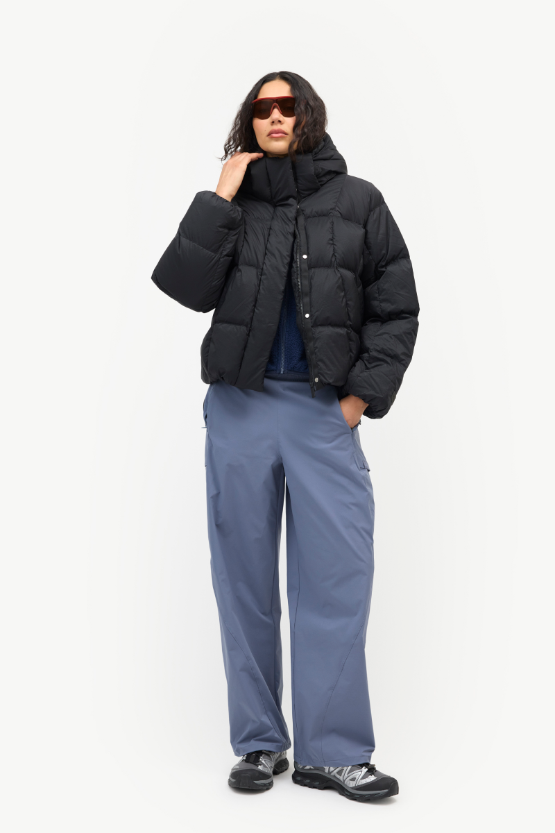 A model wearing a black puffer jacket, slate blue pants, and black sneakers, standing against a plain white studio background.