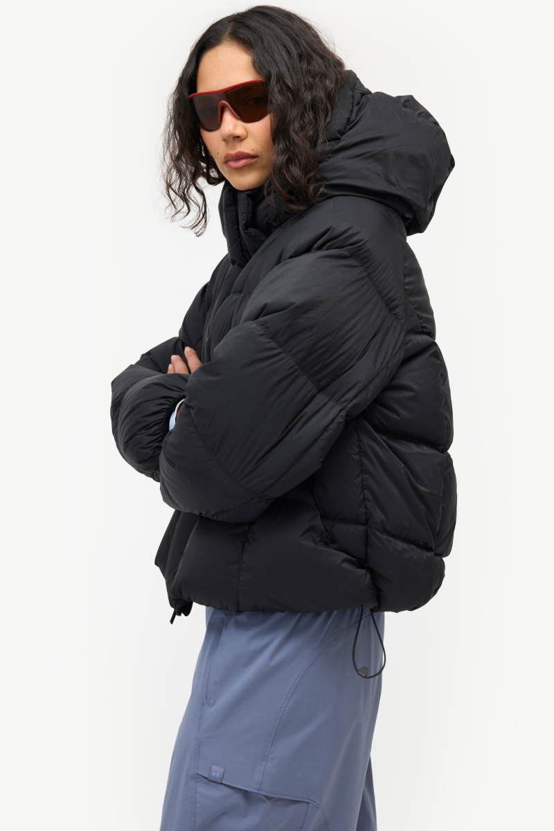 A model wearing a black puffer jacket with a hood and sunglasses, paired with slate blue pants, photographed against a clean white studio background.