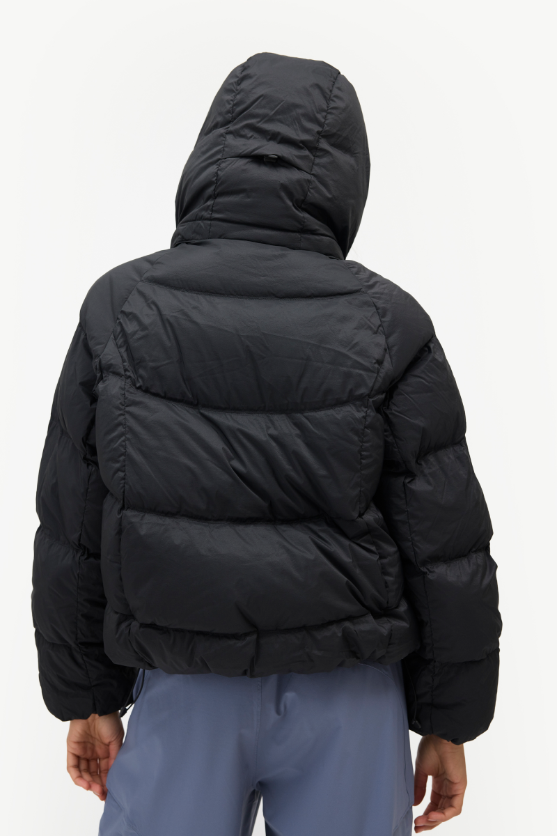 A person shown from the back wearing a black hooded puffer jacket and light blue pants against a plain light gray studio background.