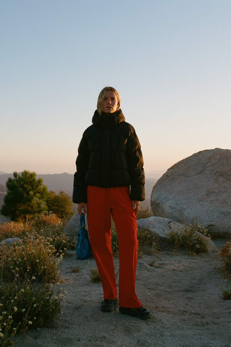 A model stands outdoors in a desert landscape at sunset, wearing a black puffer jacket and bright orange-red pants, holding a blue bag.