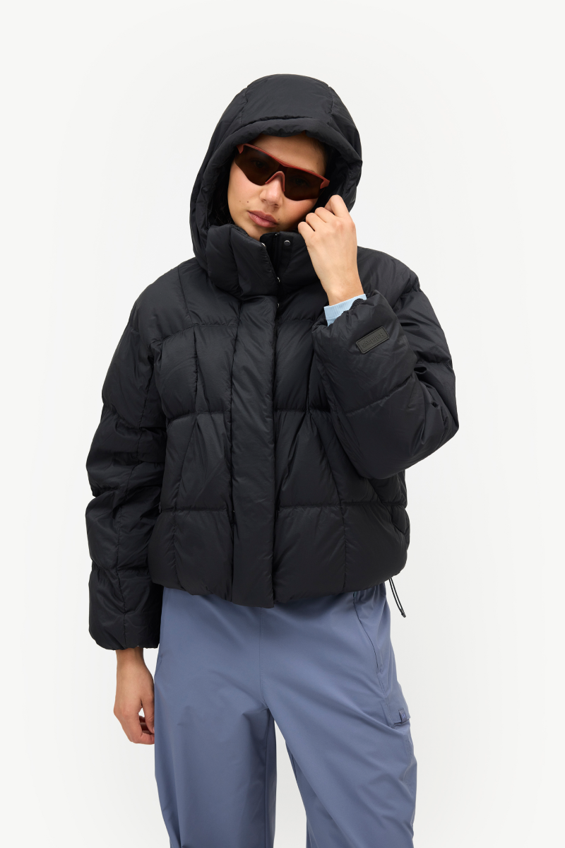 A model wearing a black cropped puffer jacket with a high collar and hood, paired with slate blue pants and sunglasses, against a plain white studio background.