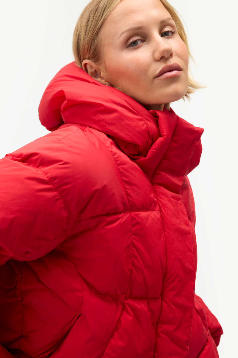 A close-up of a model wearing a bright red puffer jacket with a high padded collar, set against a clean white studio background.