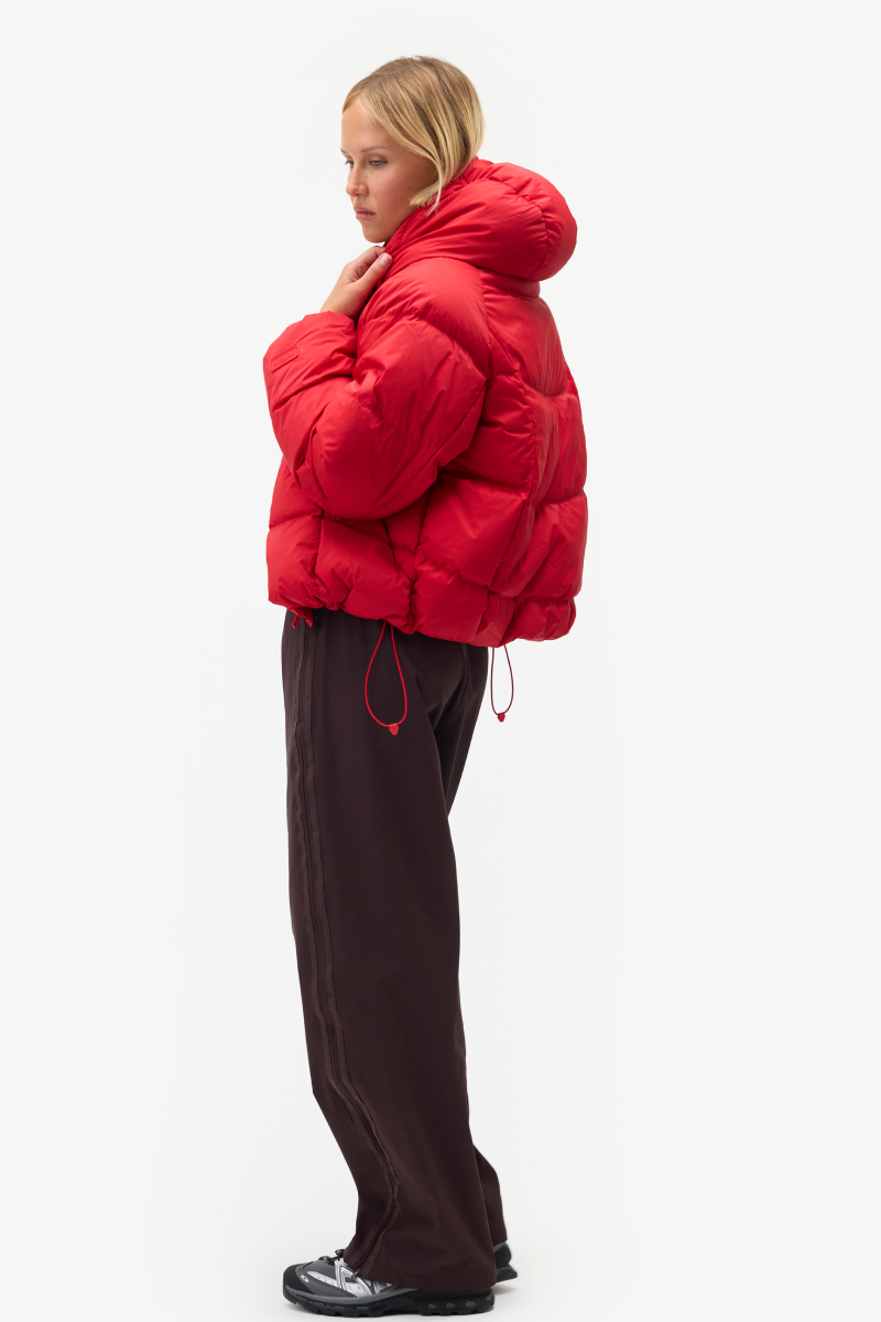 A woman stands in profile wearing a bright red puffer jacket with a high padded collar, paired with dark brown wide-leg pants and black-and-white sneakers, against a plain light gray studio background.