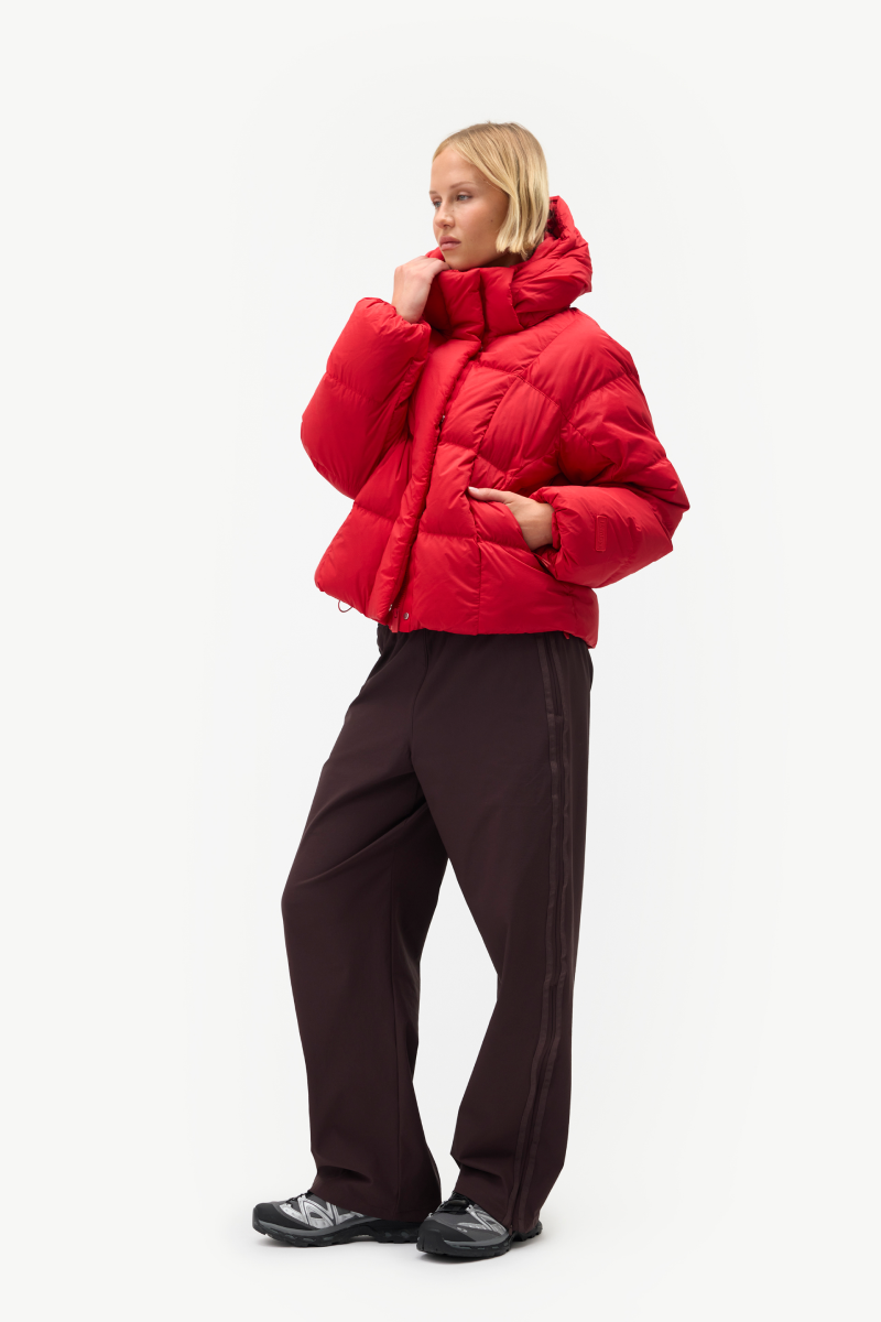 A model wearing a bright red cropped puffer jacket and dark brown wide-leg trousers, standing against a plain white studio background.