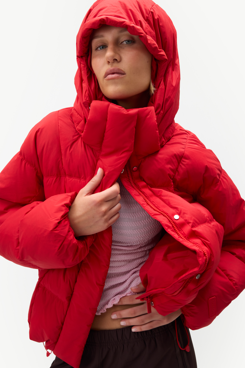 A model wearing a bright red hooded puffer jacket over a light pink ribbed top, photographed against a clean white studio background.