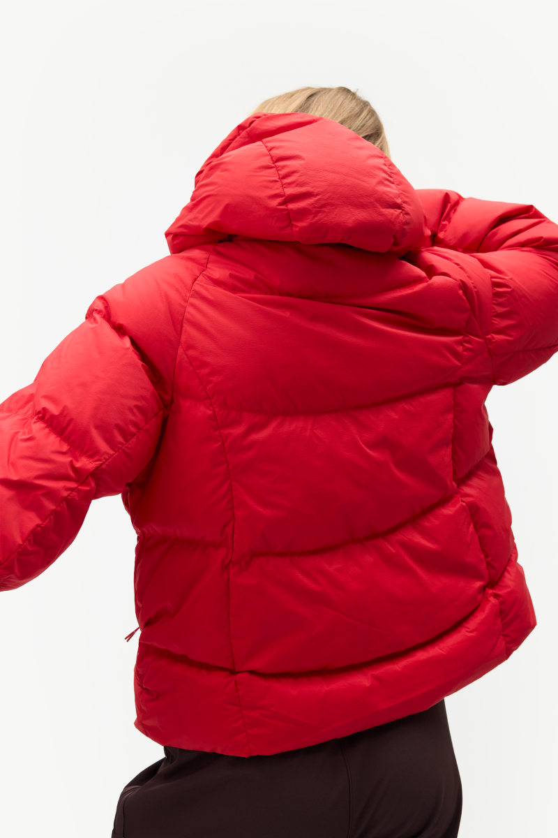 A person shown from the back wearing a bright red puffer jacket with a hood and horizontal quilted padding, against a plain light background.