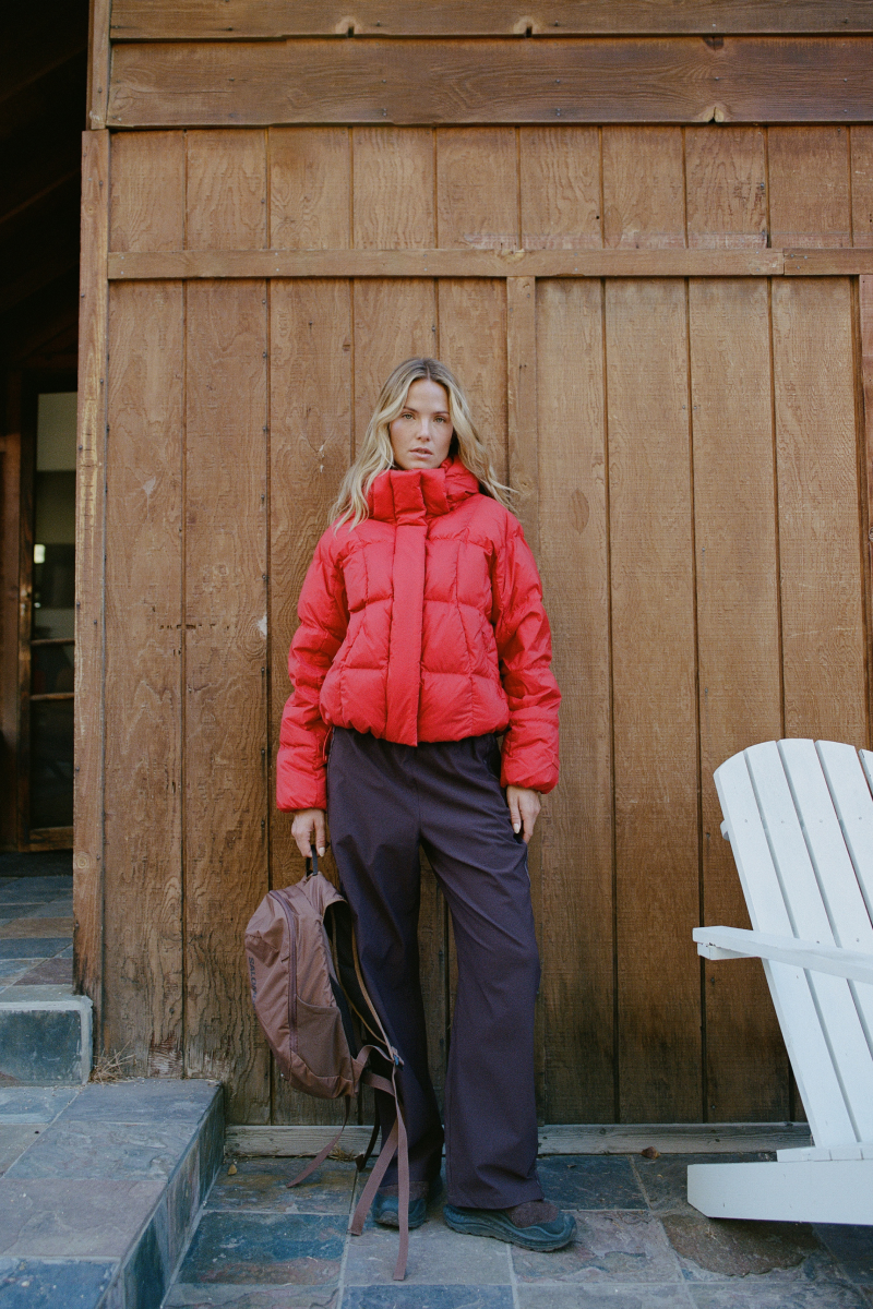 A woman stands in front of a rustic wooden wall wearing a bright red puffer jacket and dark charcoal pants, holding a brown backpack. A white Adirondack chair is visible on the right.