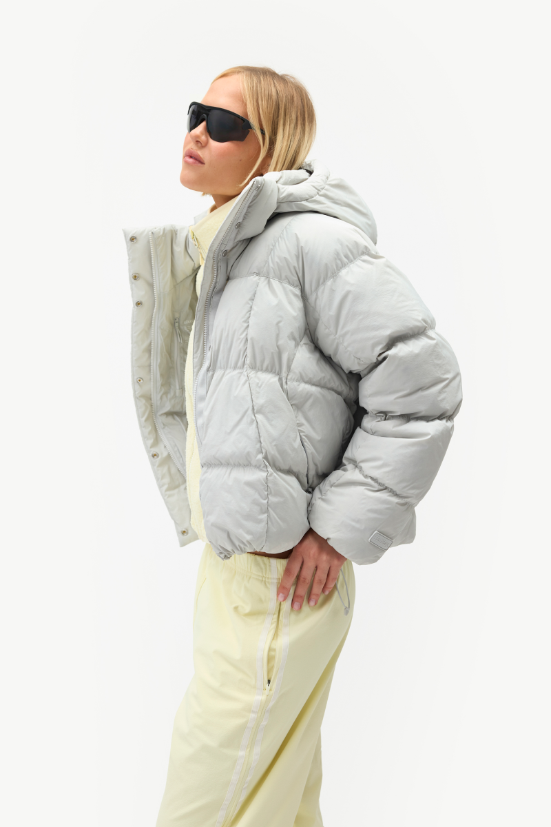 A model wearing a light gray puffer jacket, cream pants, and black sunglasses, shown in profile against a plain white studio background.