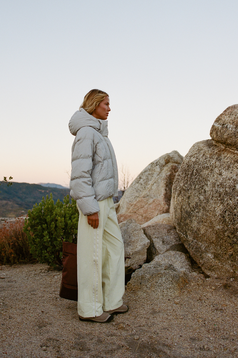 A woman stands outdoors in a rocky desert setting at dusk, wearing a light gray puffer jacket, cream wide-leg pants, and brown slip-on shoes.