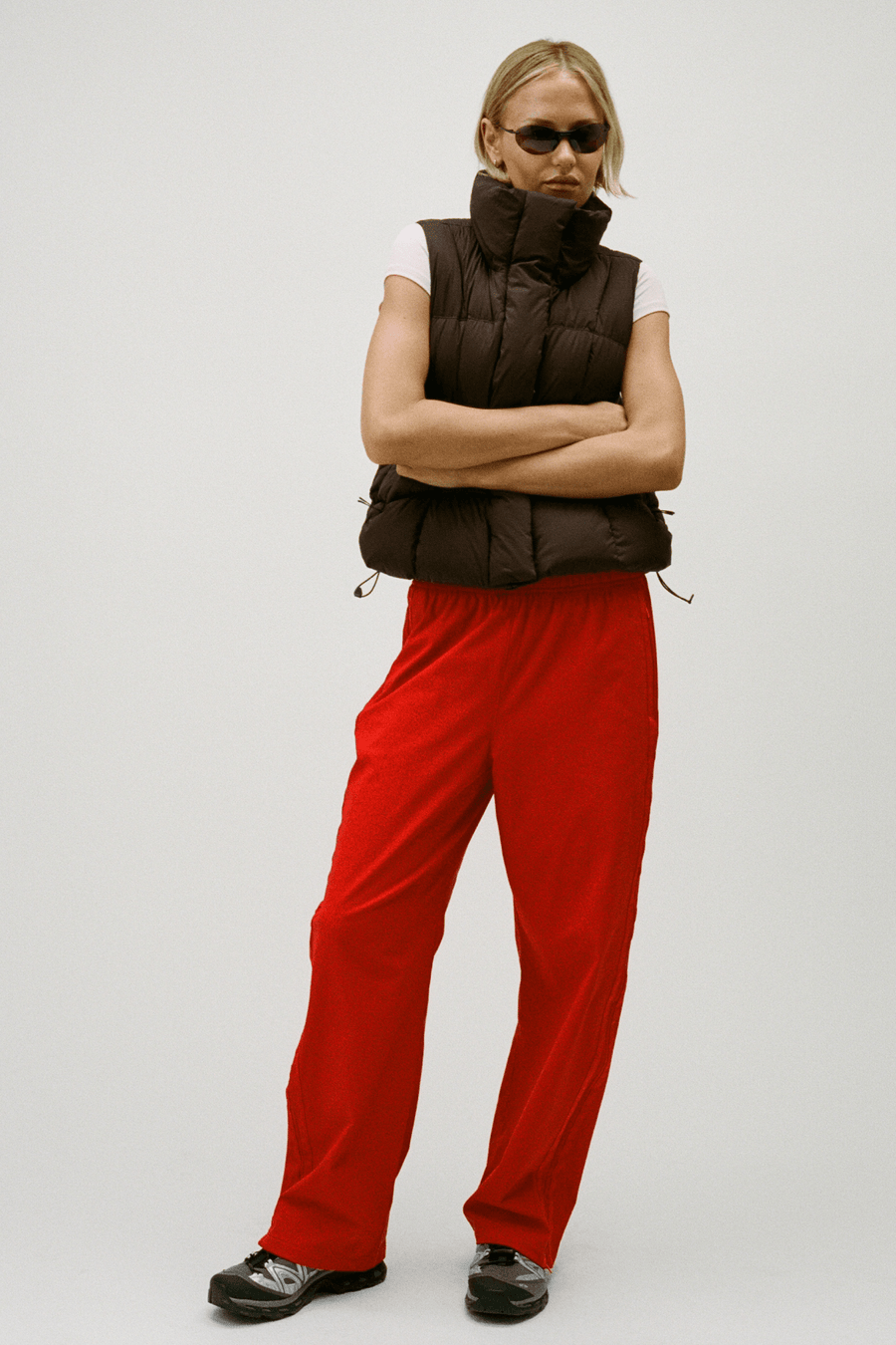 A model wearing a dark brown puffer vest, bright red pants, a white sleeveless top, and black shoes, standing against a light gray studio background.