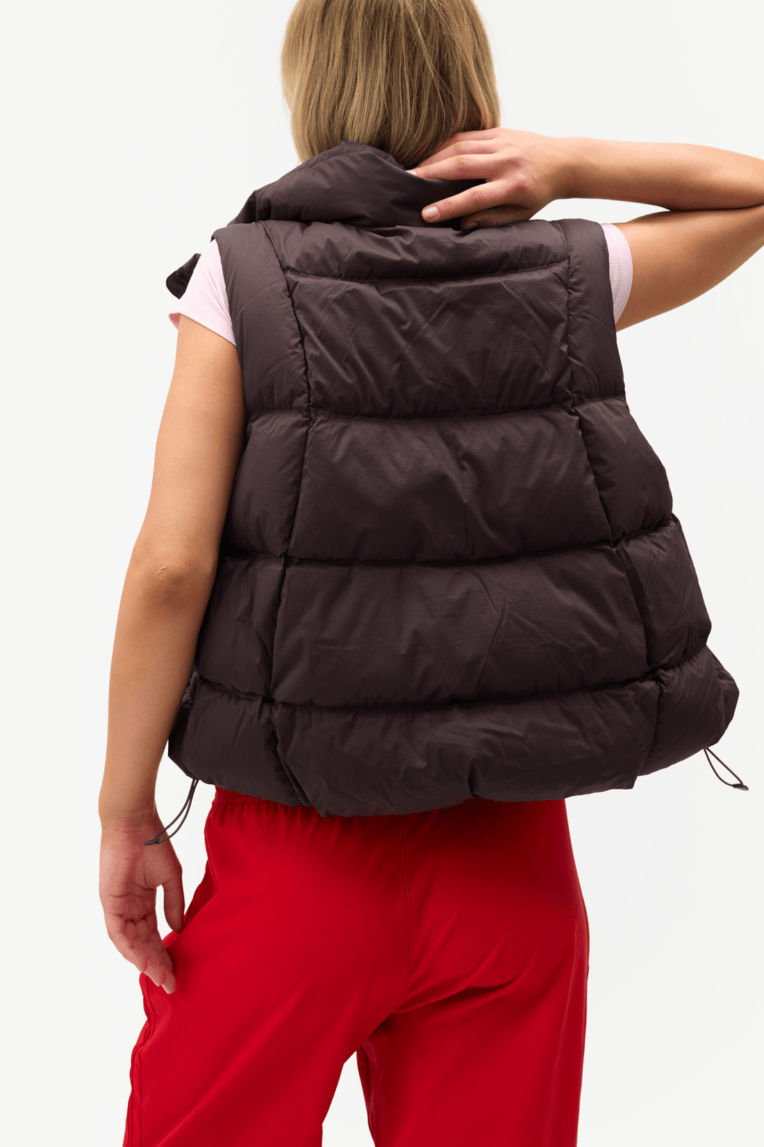 A woman shown from the back wearing a dark brown puffer vest over a light pink sleeveless top and bright red pants, against a plain white studio background.