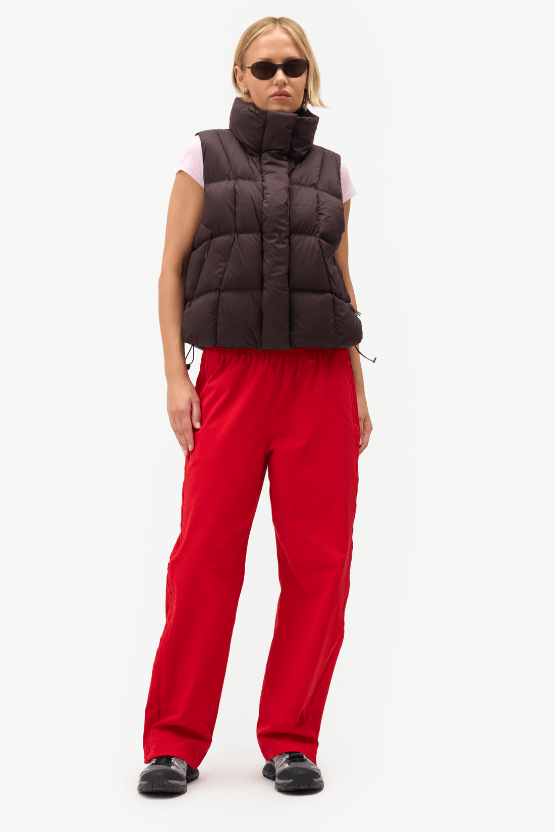 A model wearing a dark brown puffer vest over a light pink sleeveless top, paired with bright red pants and black shoes, standing against a plain white studio background.