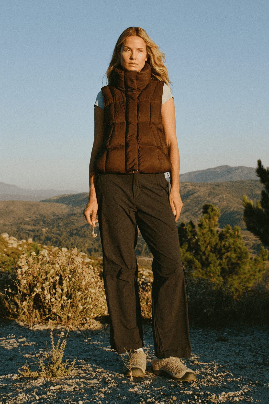 A woman stands outdoors in a mountain landscape wearing a brown sleeveless puffer vest over a light gray top, dark wide-leg pants, and hiking boots.
