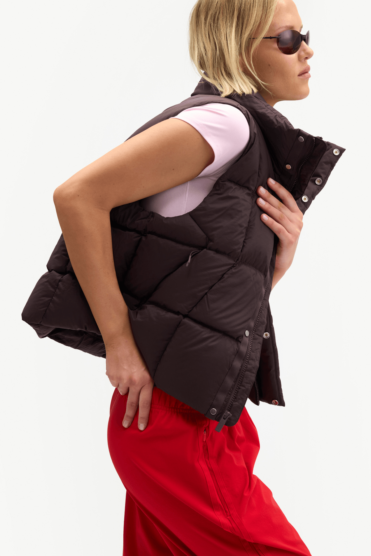 A woman wearing a dark brown puffer vest over a light pink top and bright red pants, shown from the side against a plain white studio background.