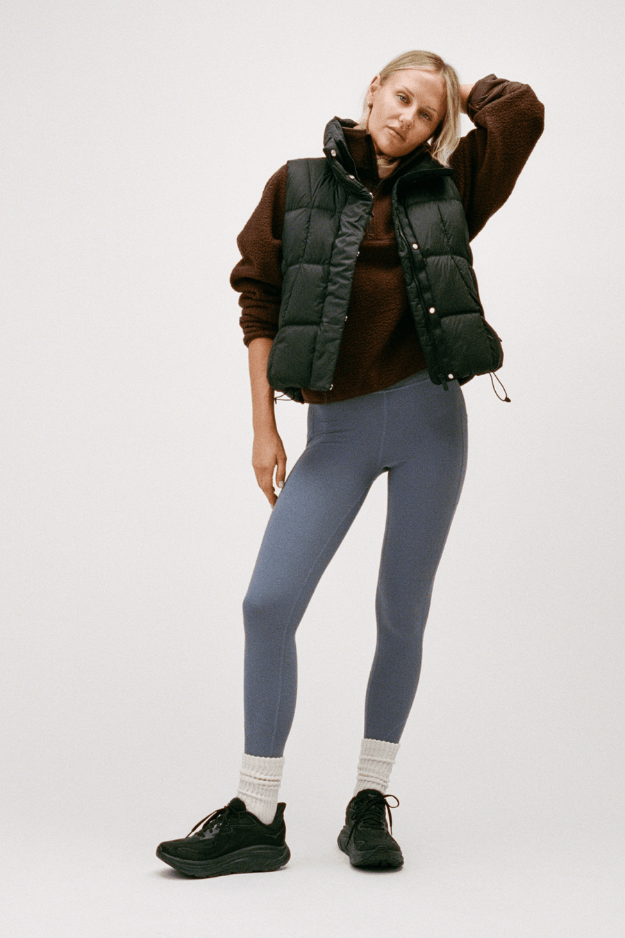 A woman modeling a black puffer vest over a brown long-sleeve top, paired with slate blue leggings, white socks, and black sneakers, against a plain light gray studio background.
