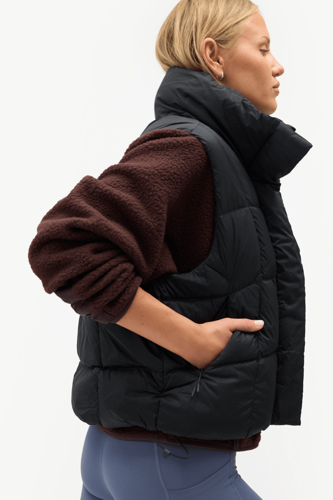 A woman shown in side profile wearing a black quilted puffer vest over a dark brown sherpa long-sleeve top and blue leggings, against a plain white studio background.
