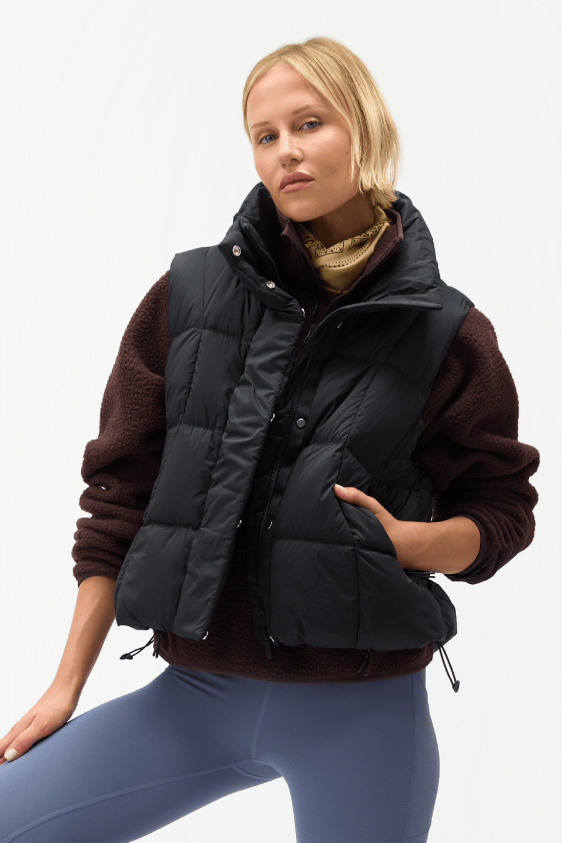A woman wearing a black puffer vest over a brown fleece sweater and blue leggings, posed against a clean white studio background.