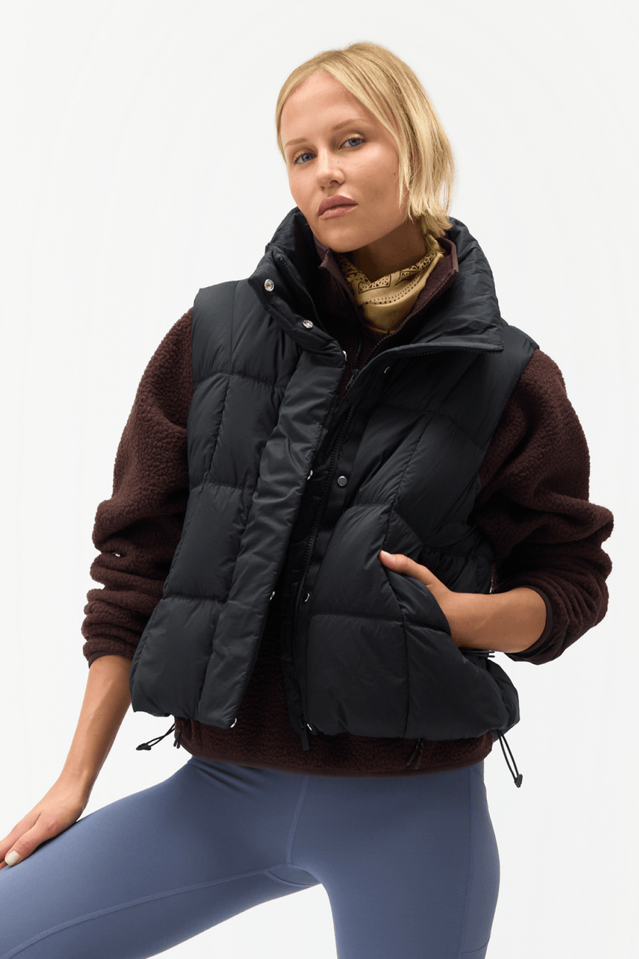 A woman wearing a black puffer vest over a brown fleece sweater and blue leggings, posed against a clean white studio background.