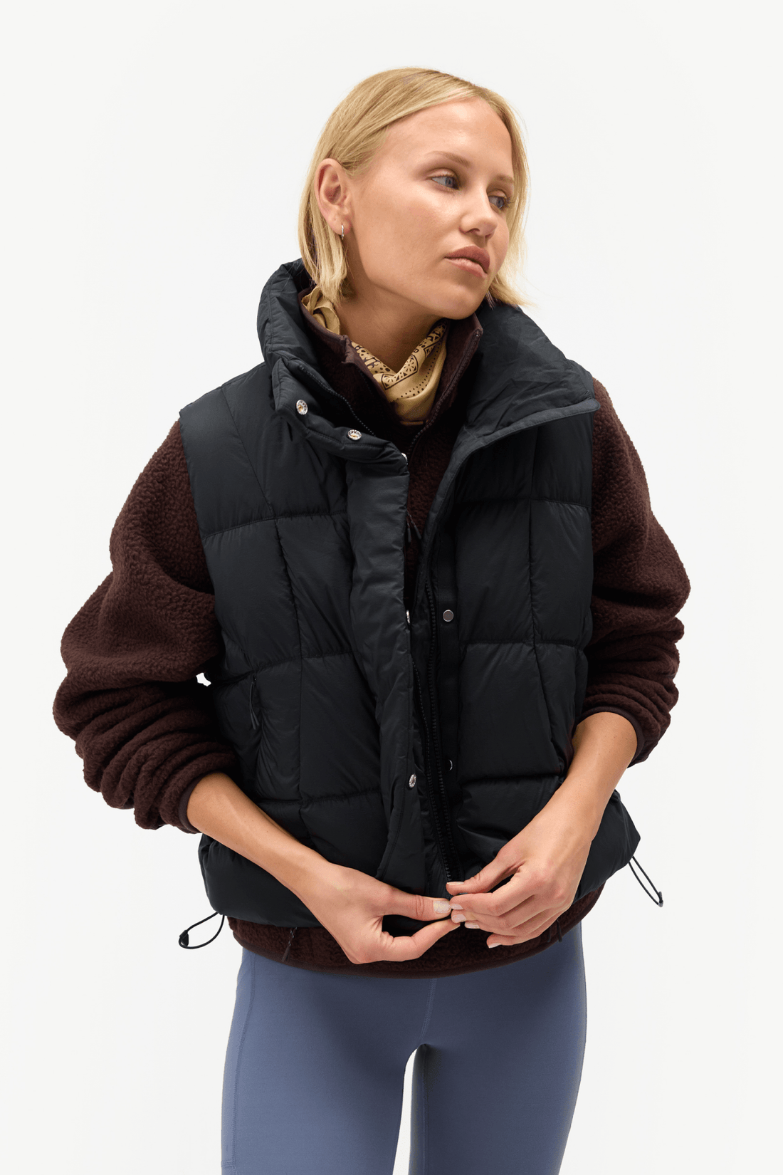A woman wearing a black puffer vest over a dark brown fleece sweater and blue leggings, photographed against a clean white studio background.