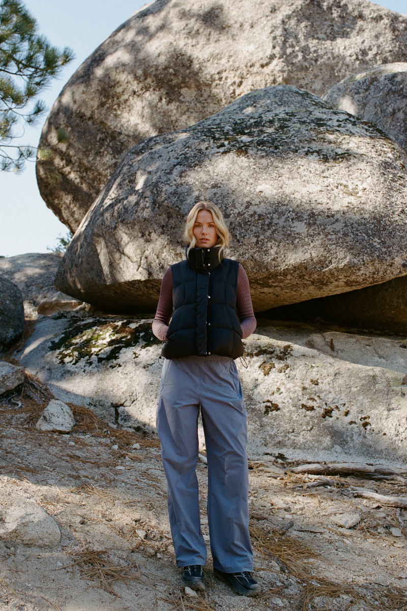 A woman stands outdoors in front of large rock formations, wearing a black quilted sleeveless puffer vest over a mauve-brown long-sleeve top, loose light blue-gray pants, and dark sneakers.