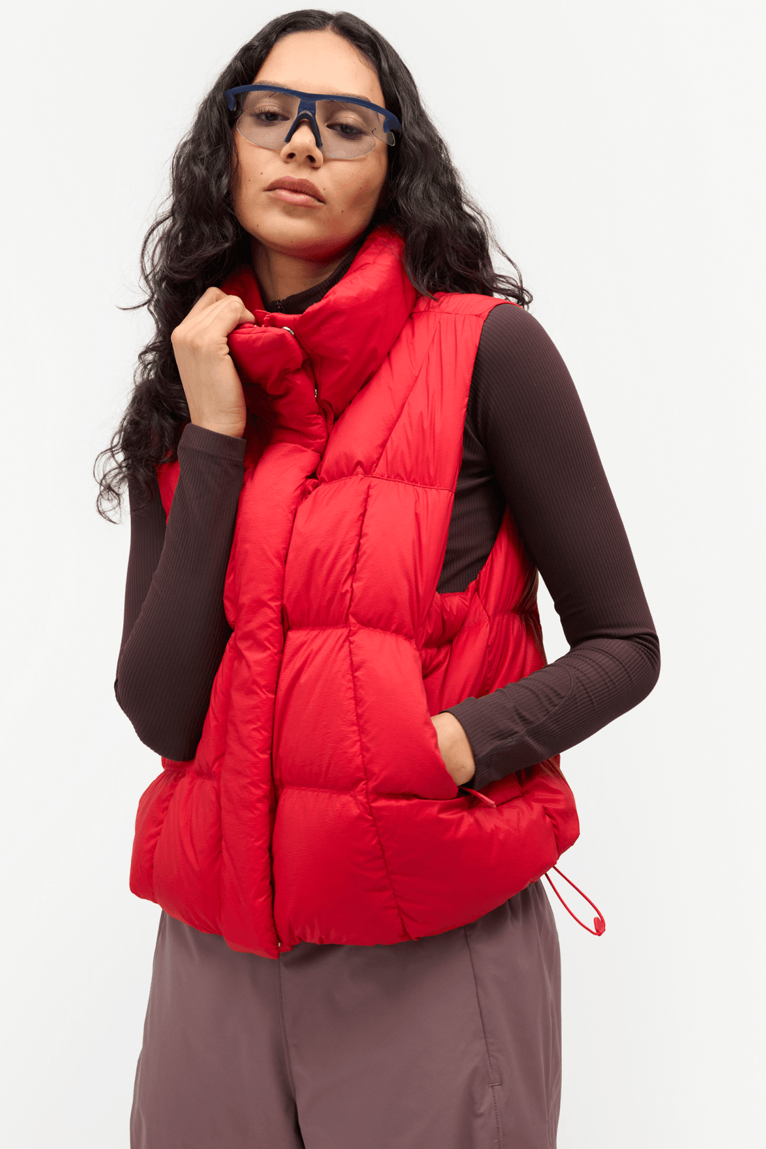 A woman wearing a bright red puffer vest over a dark brown long-sleeve top and mauve pants, photographed against a clean white studio background.