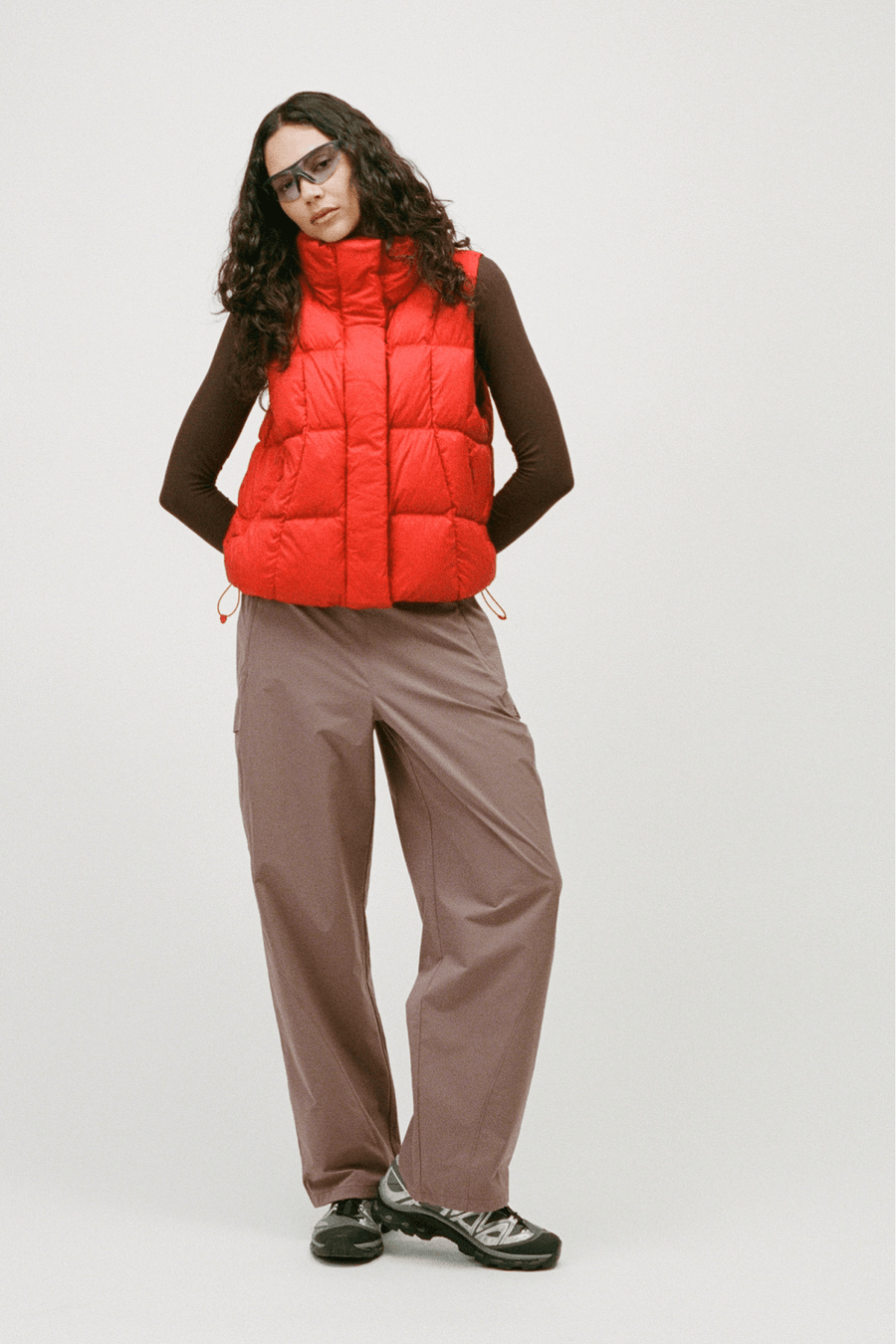 A model wearing a bright red puffer vest over a dark brown long-sleeve top, paired with wide-leg taupe pants and black sneakers, standing against a light gray studio background.