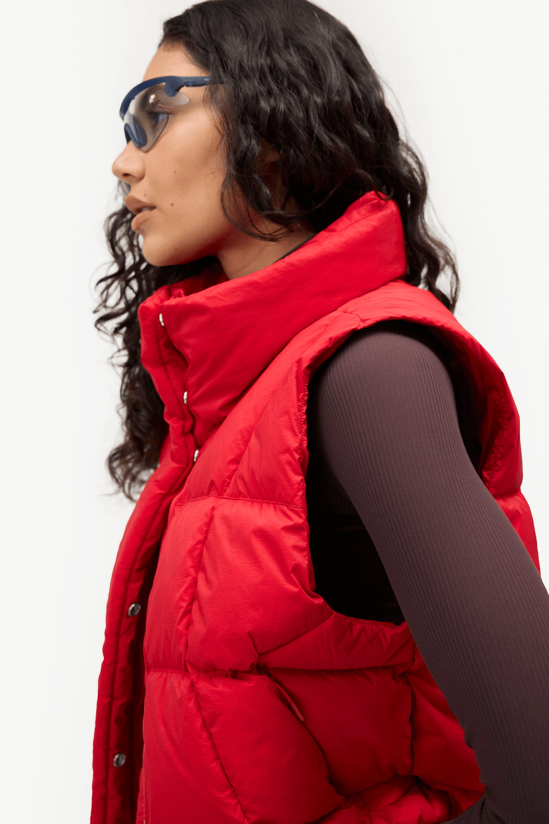 A woman in profile wearing a bright red puffer vest over a dark ribbed long-sleeve top, with blue-tinted sunglasses, against a plain light background.