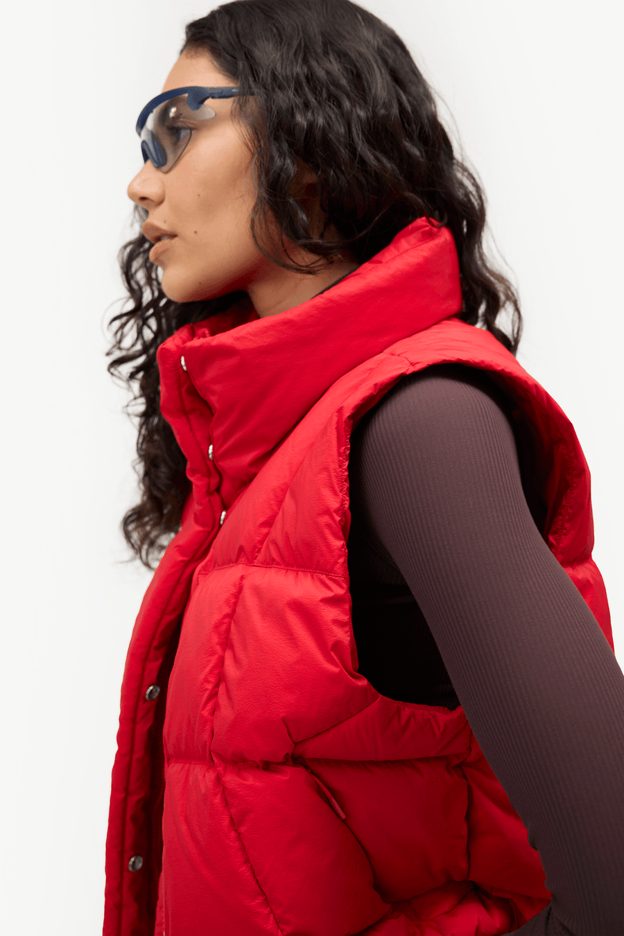 A woman in profile wearing a bright red puffer vest over a dark ribbed long-sleeve top, with blue-tinted sunglasses, against a plain light background.