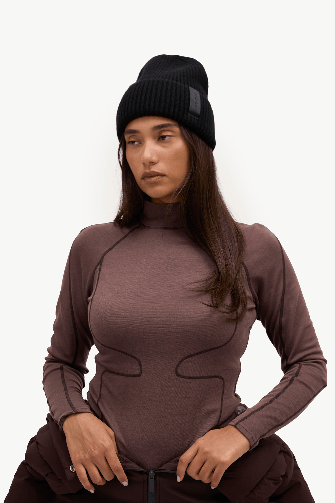 A woman wearing a fitted long-sleeve athletic top in a muted mauve-brown color, paired with dark brown pants and a black knit beanie, posed against a clean white studio background.