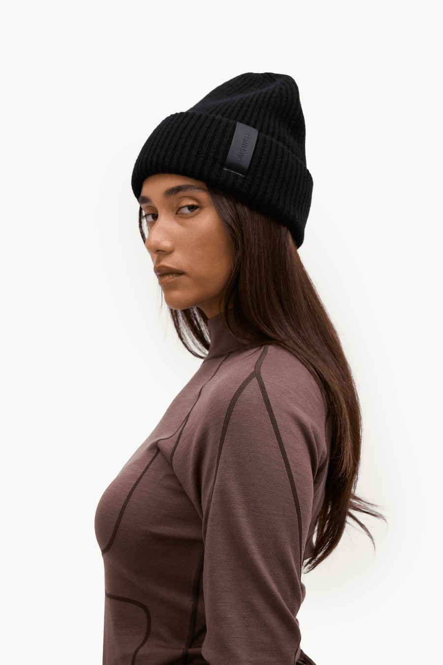 A woman in profile wearing a black ribbed knit beanie with a small rectangular black patch on the side, paired with a brown athletic top, against a plain white background.