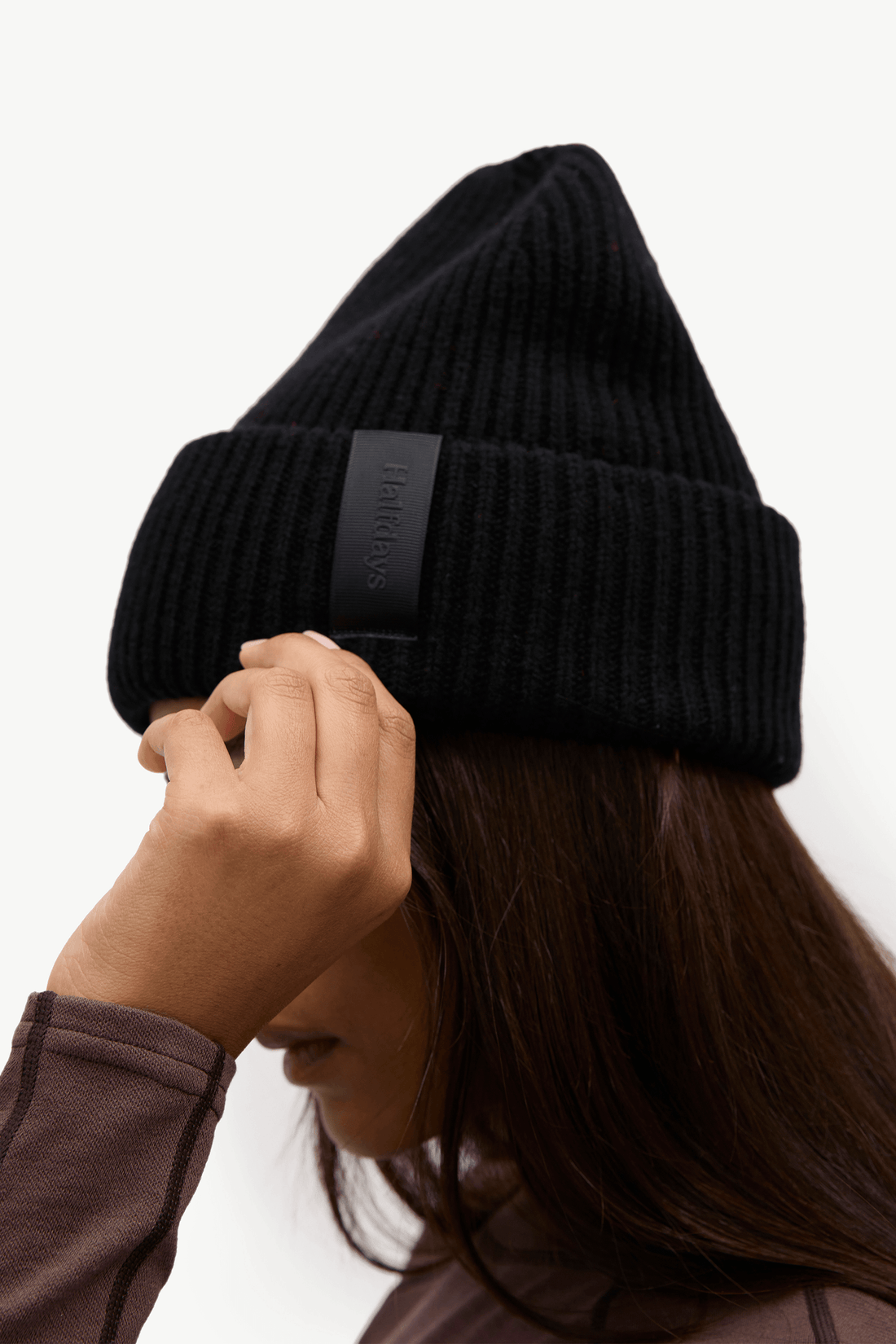 A close-up of a person wearing a black ribbed knit beanie with a black fabric brand tab on the cuff, against a plain light background.