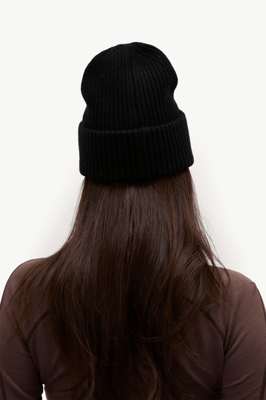 A person viewed from the back wearing a black ribbed knit beanie hat, with long dark brown hair and a brown top against a plain white background.