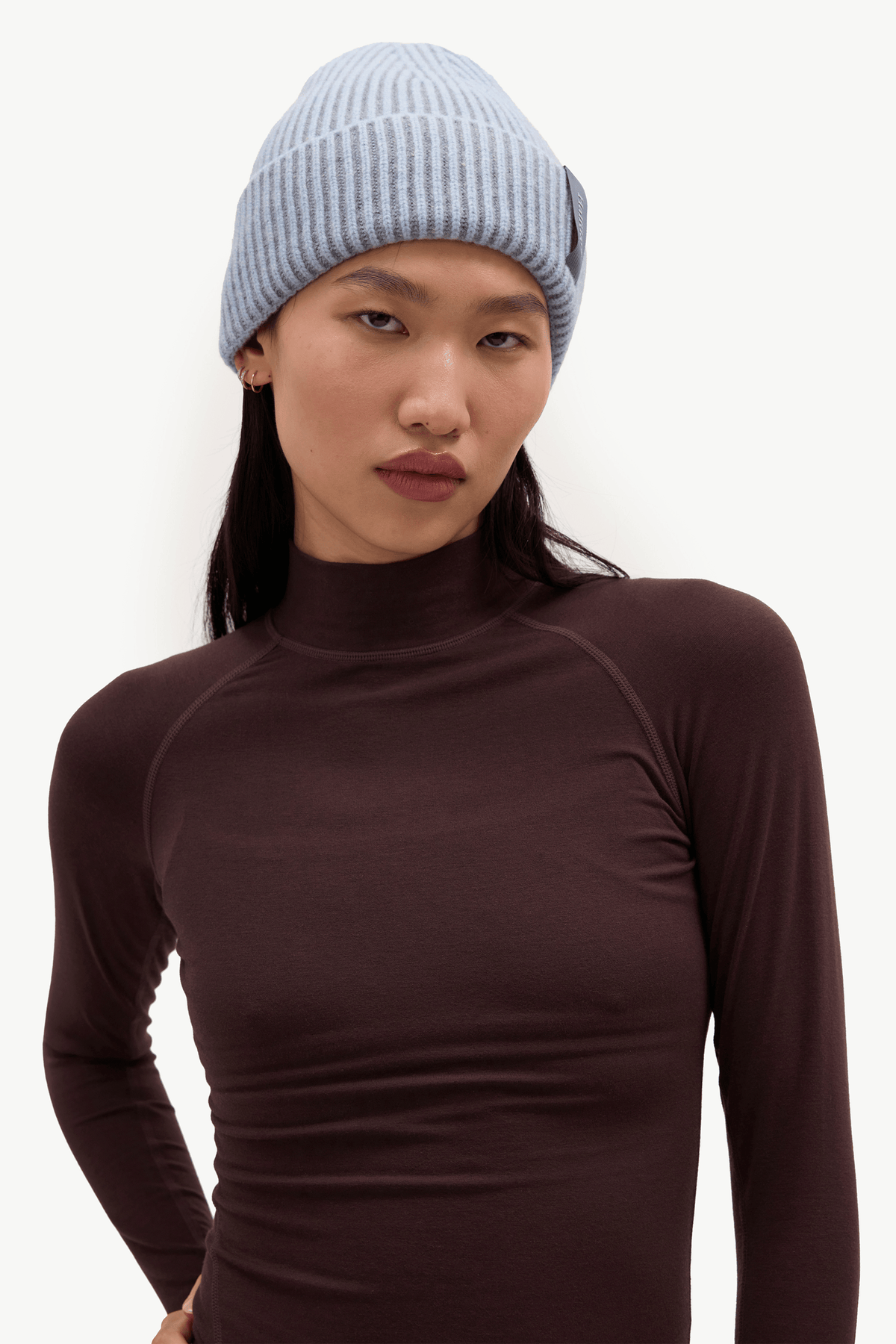 A model wearing a light blue ribbed knit beanie and a dark brown long-sleeve mock neck top, photographed against a clean white studio background.