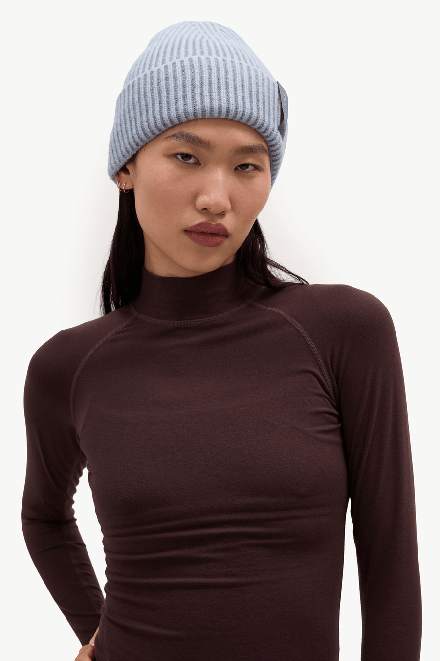 A model wearing a light blue ribbed knit beanie and a dark brown long-sleeve mock neck top, photographed against a clean white studio background.