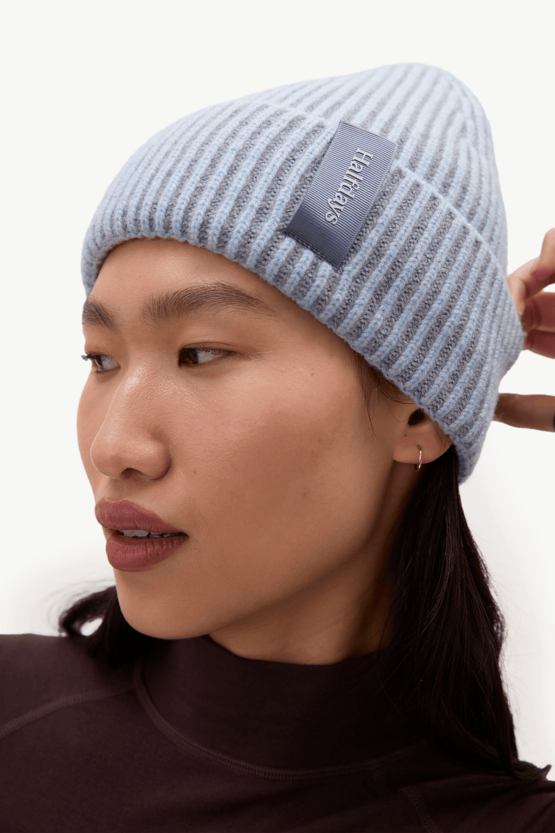 A close-up of a model wearing a light blue and white striped knit beanie with a dark gray brand patch that reads 'Halfdays'.