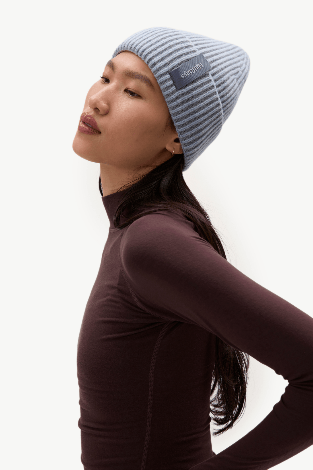 A woman wearing a light blue and gray striped knit beanie with a dark gray brand patch that reads 'Halfdays', paired with a dark plum long-sleeve mock neck top, photographed against a clean white background.