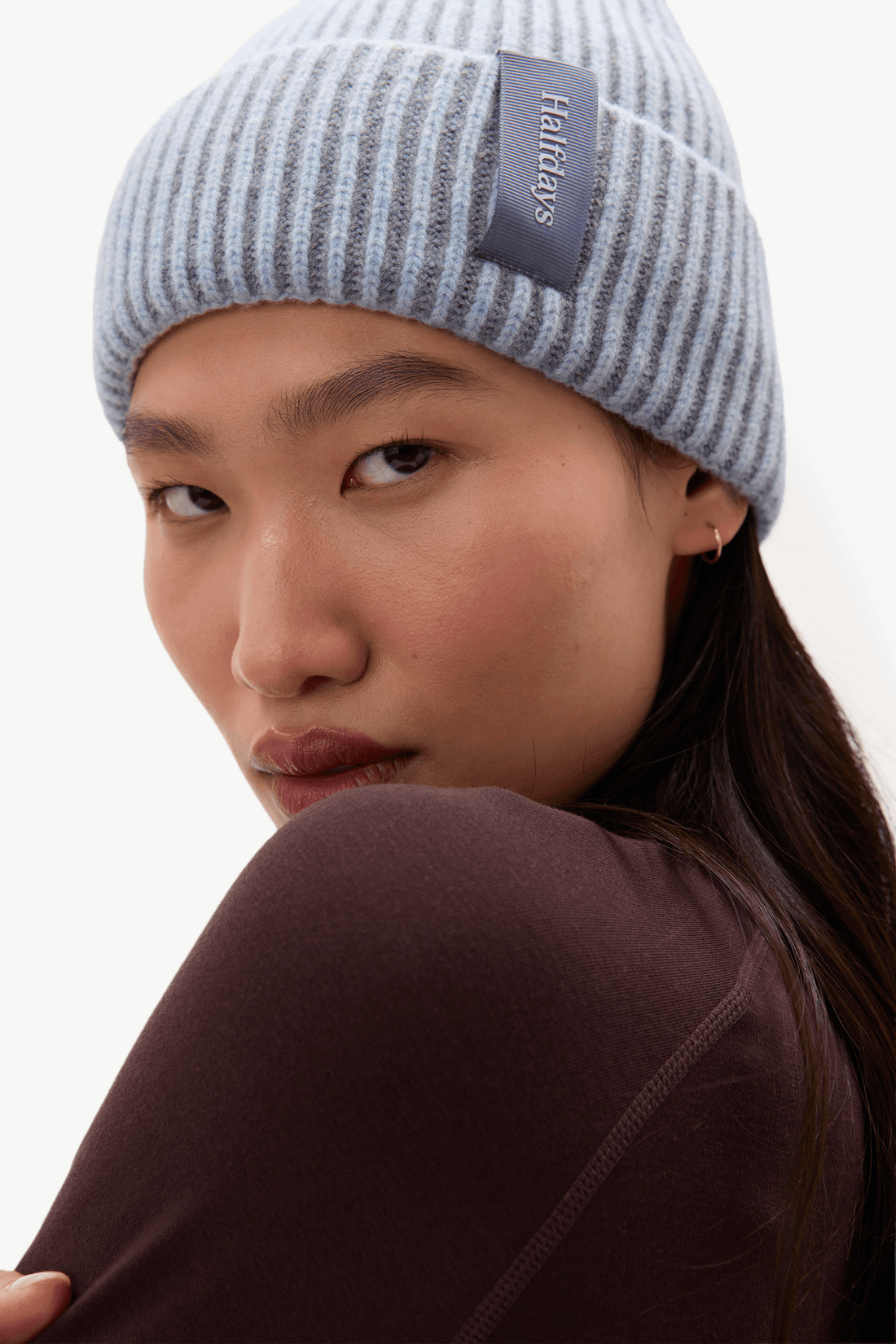 A close-up of a model wearing a light gray and white striped ribbed beanie with a dark gray brand patch that reads 'Halfdays', paired with a dark brown top against a plain white background.