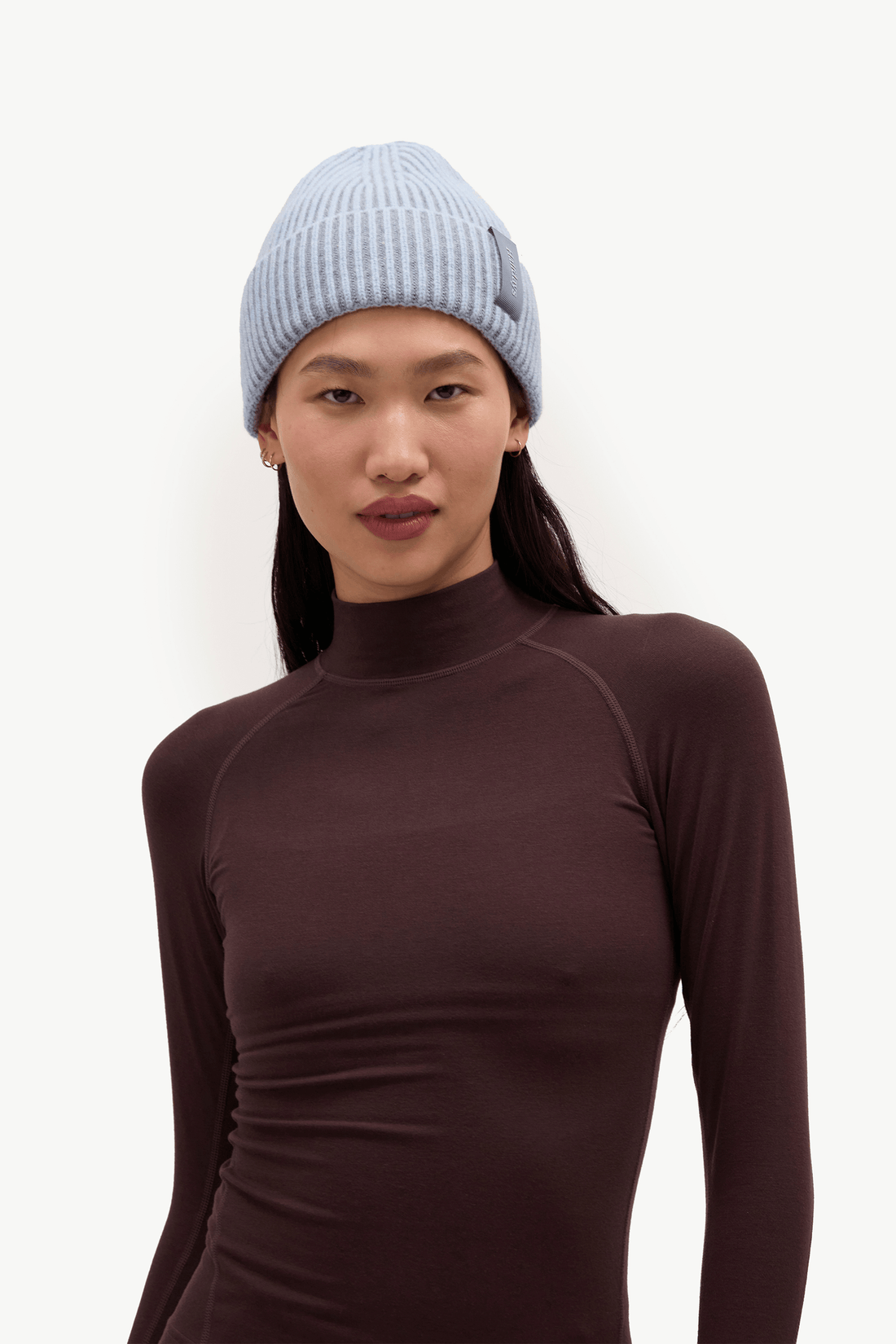 A woman wearing a light blue ribbed knit beanie and a dark brown long-sleeve mock neck top, photographed against a plain white background.